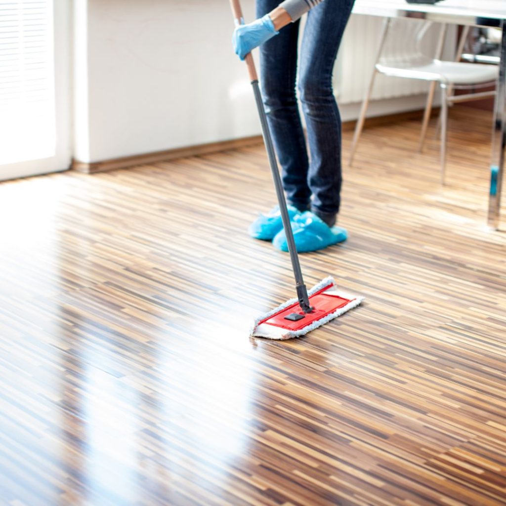 Person mopping a polished hardwood floor in a clean, modern interior while wearing blue shoe covers and gloves.